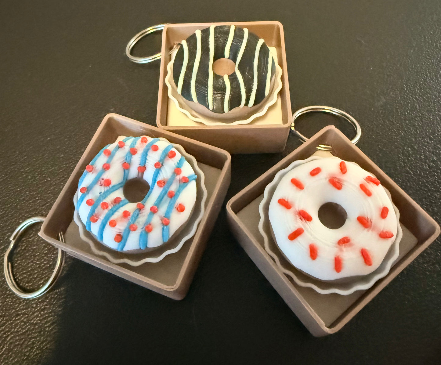 Three donut-shaped keychains in decorative boxes on a dark surface